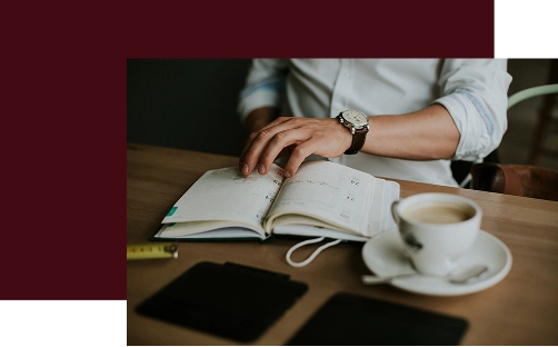 Osoba ubrana w białą koszulę siedzi przy drewnianym stole i czyta otwarty terminarz. Obok leży zegarek na rękę, filiżanka kawy i tablet, tworząc atmosferę skupienia i relaksu.