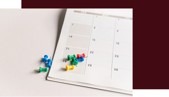 Na powierzchni leży strona kalendarza, pokryta kolorowymi pinezkami w kolorach niebieskim, zielonym, żółtym i czerwonym. Obraz ten symbolizuje planowanie i organizację.