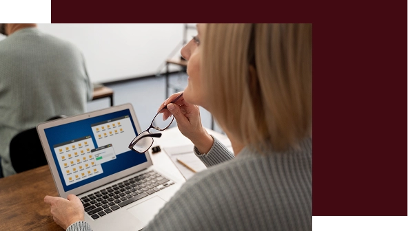 Kobieta w swetrze trzyma okulary i patrzy na laptopa, na którego ekranie wyświetlane są foldery. Siedzi przy drewnianym biurku, sprawiając wrażenie skupionej i zamyślonej.